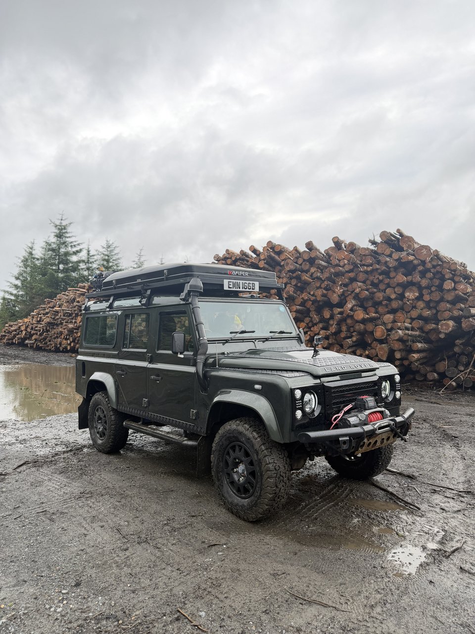 All Terrain Academy’s green Land Rover Defender 110 beside stacked logs on a muddy forest track