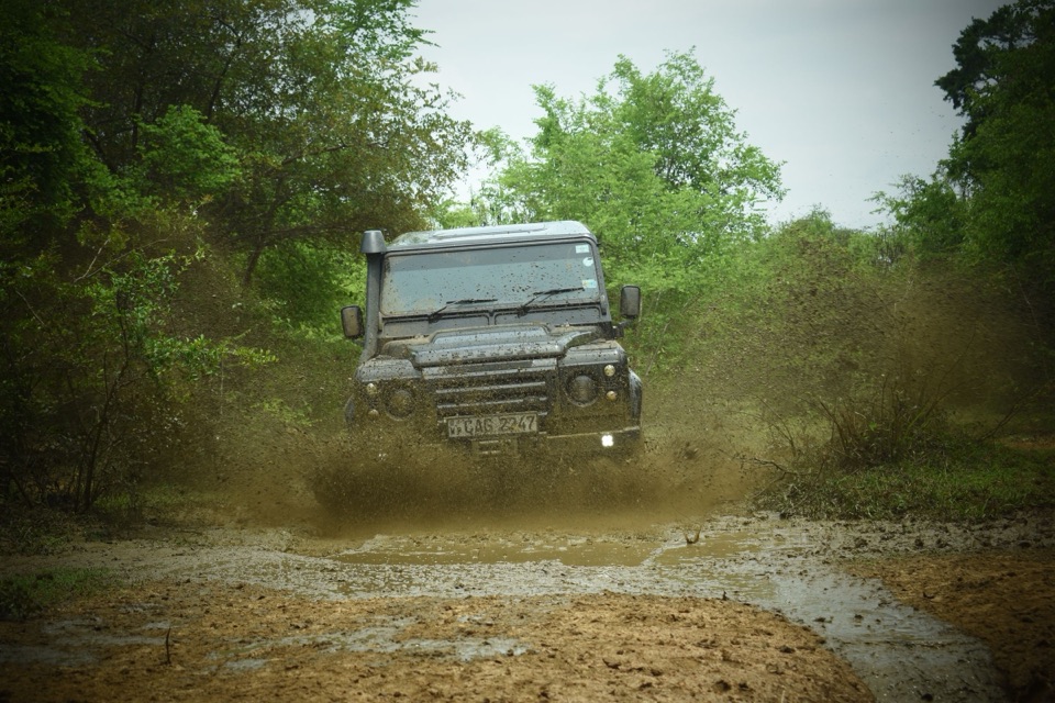 A Land Rover Defender coming head-on through a muddy woodland track, water spraying from the front wheels