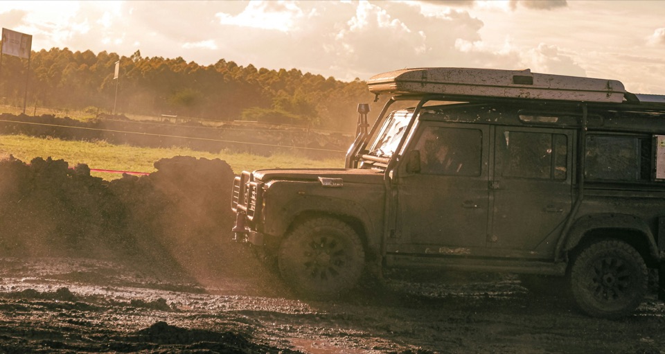 A Land Rover Defender 110 with a roof tent driving along a dirt track at dusk, dust lit by the low sun