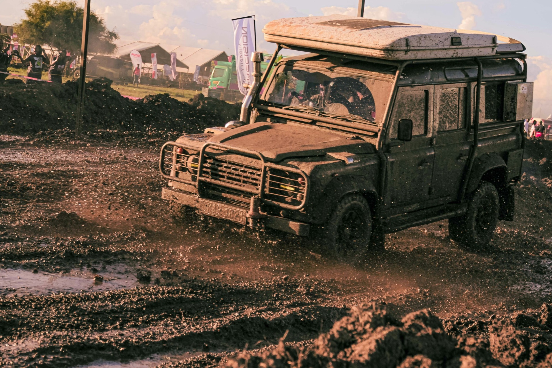 A dark green Land Rover Defender 110 with a roof tent driving through a mud pool, shot low from the front as the wheels throw up mud