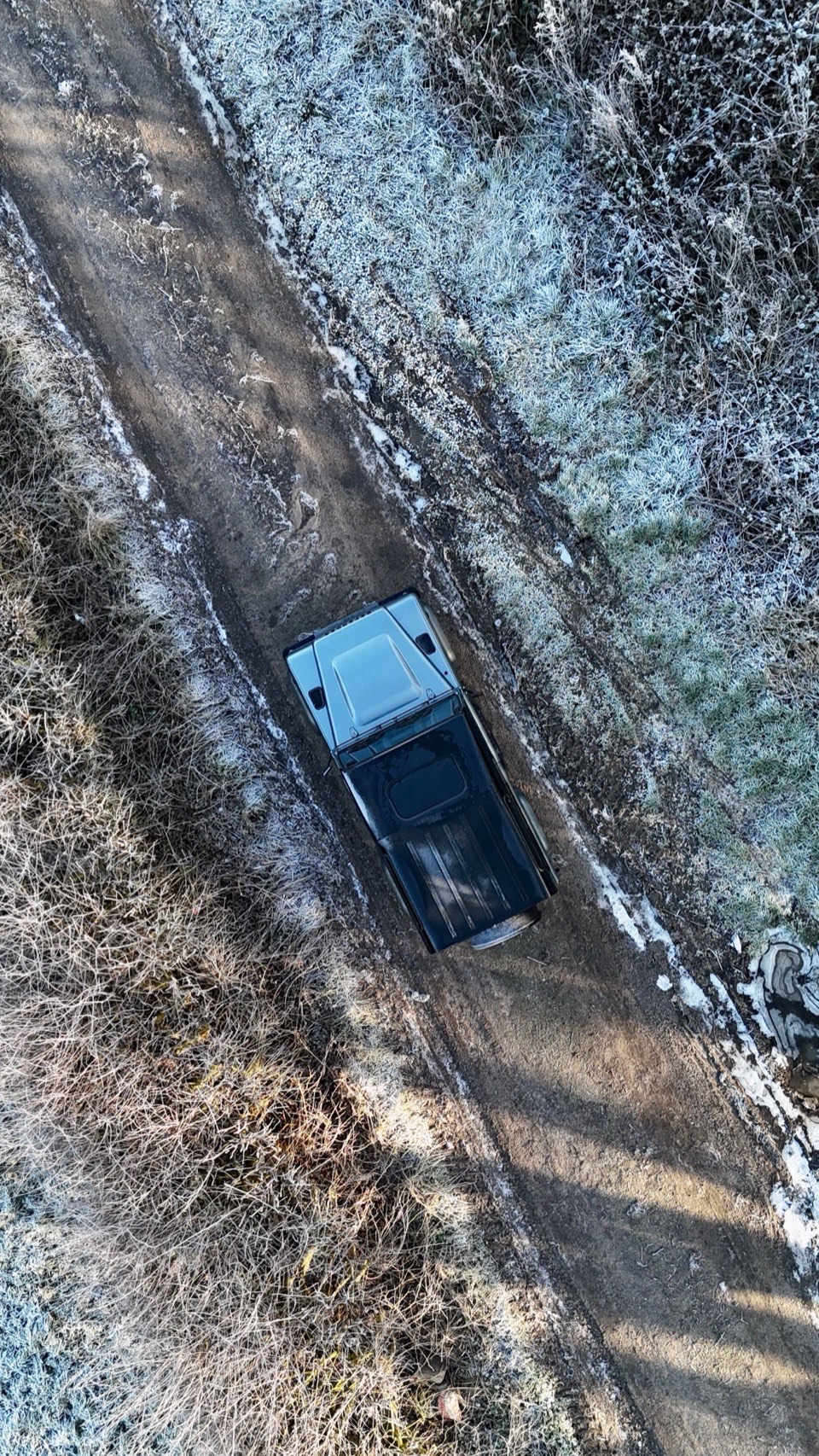A Land Rover Defender seen from directly above on a frosty dirt track between white grass verges